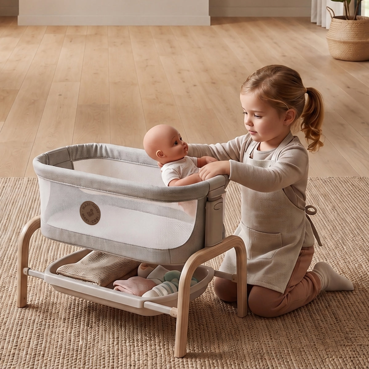 Young child placing a baby doll into the Maxi-Cosi Iora toy bassinet on the living room floor.