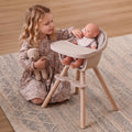 Young child seated on a rug feeding an 18-inch baby doll in the Maxi-Cosi Moa toy high chair.